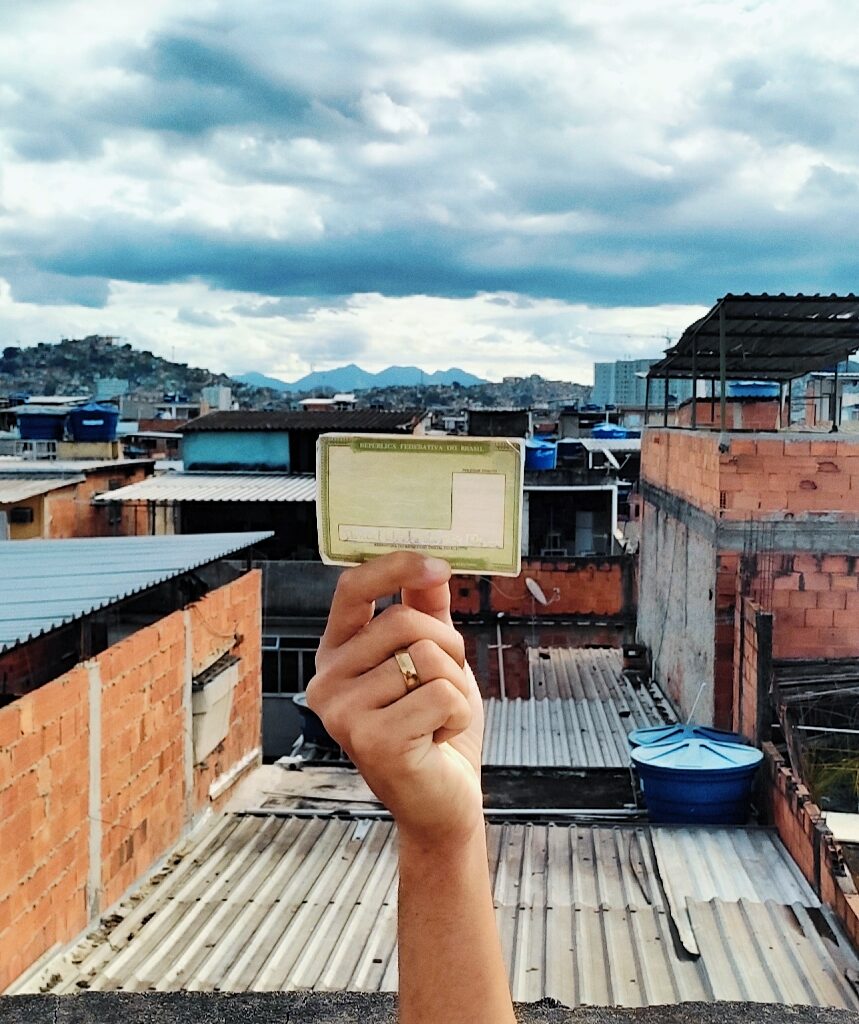 Mão jovem segura título de eleitor para o alto; ao fundo, casas de uma das favelas da Maré
