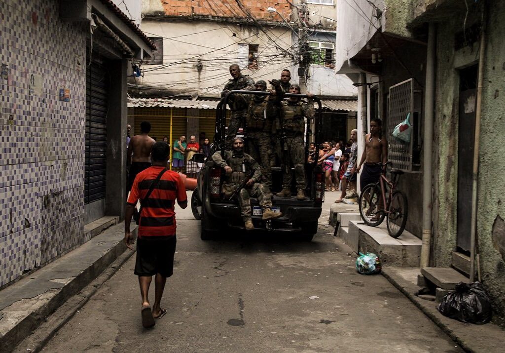 Homem com camisa do Flamengo andando em rua com carro repleto de militares armados durante operação na Baixa do Sapateiro