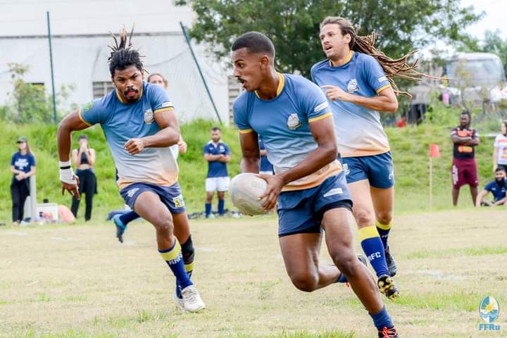 Homem jovem segura bola de rugby com dois outros jogadores em segundo plano